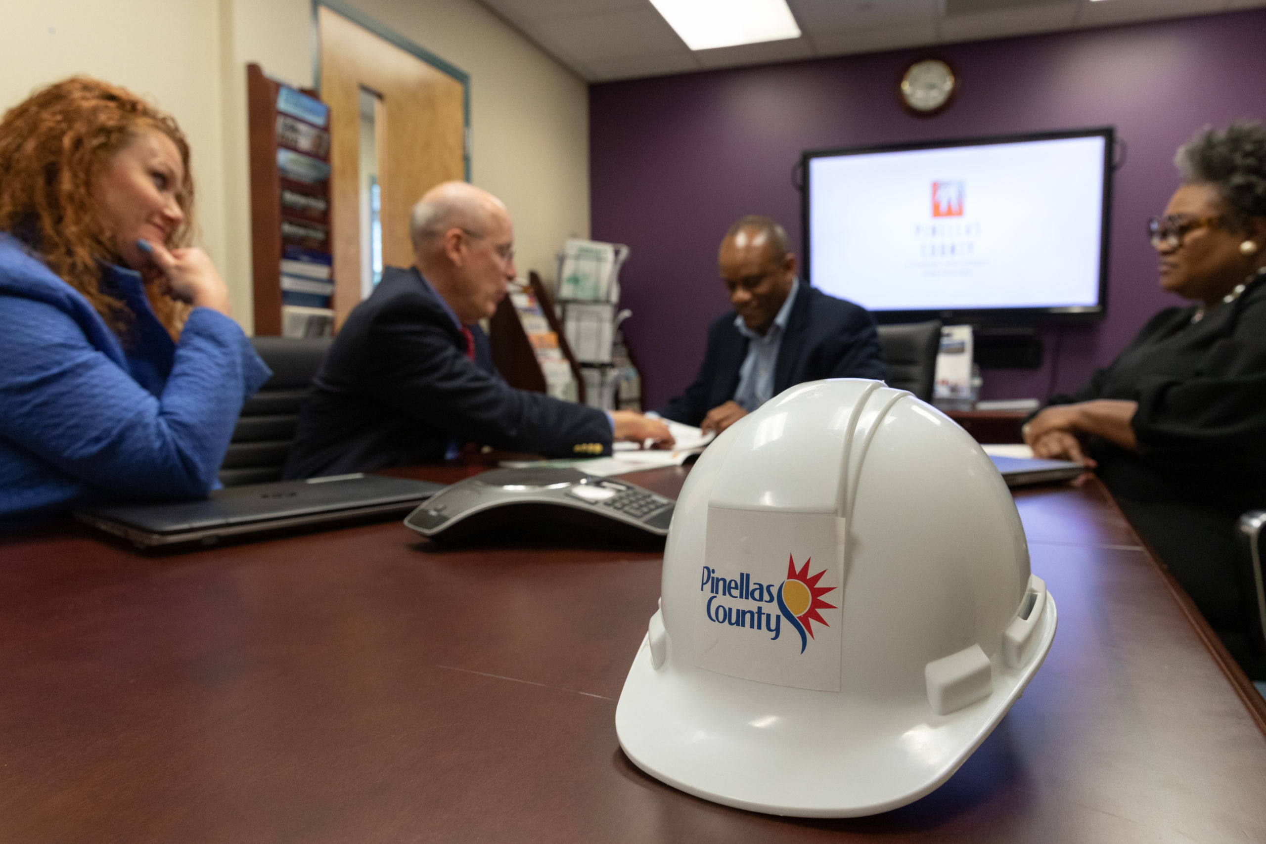 People sitting at a table discussing business and government contracting, and hard hat with the Pinellas County logo is on the table.