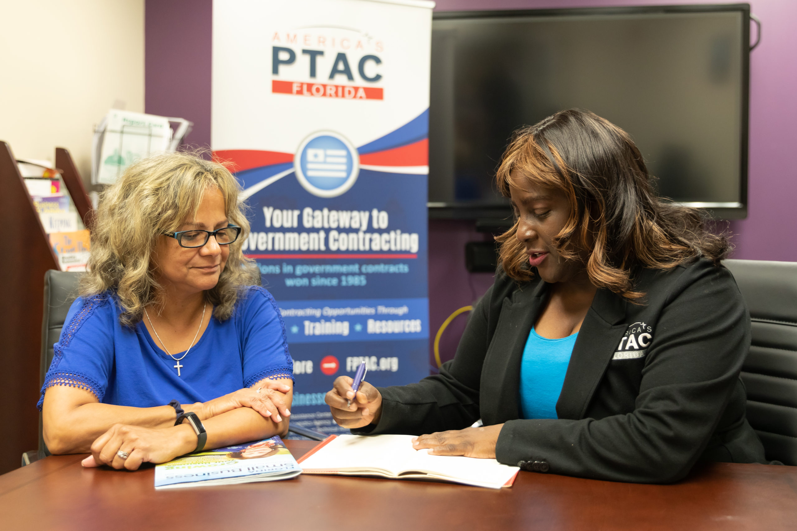 Image of two women meeting at a table with a sign behind them for the Procurement Technical Assistance Program.