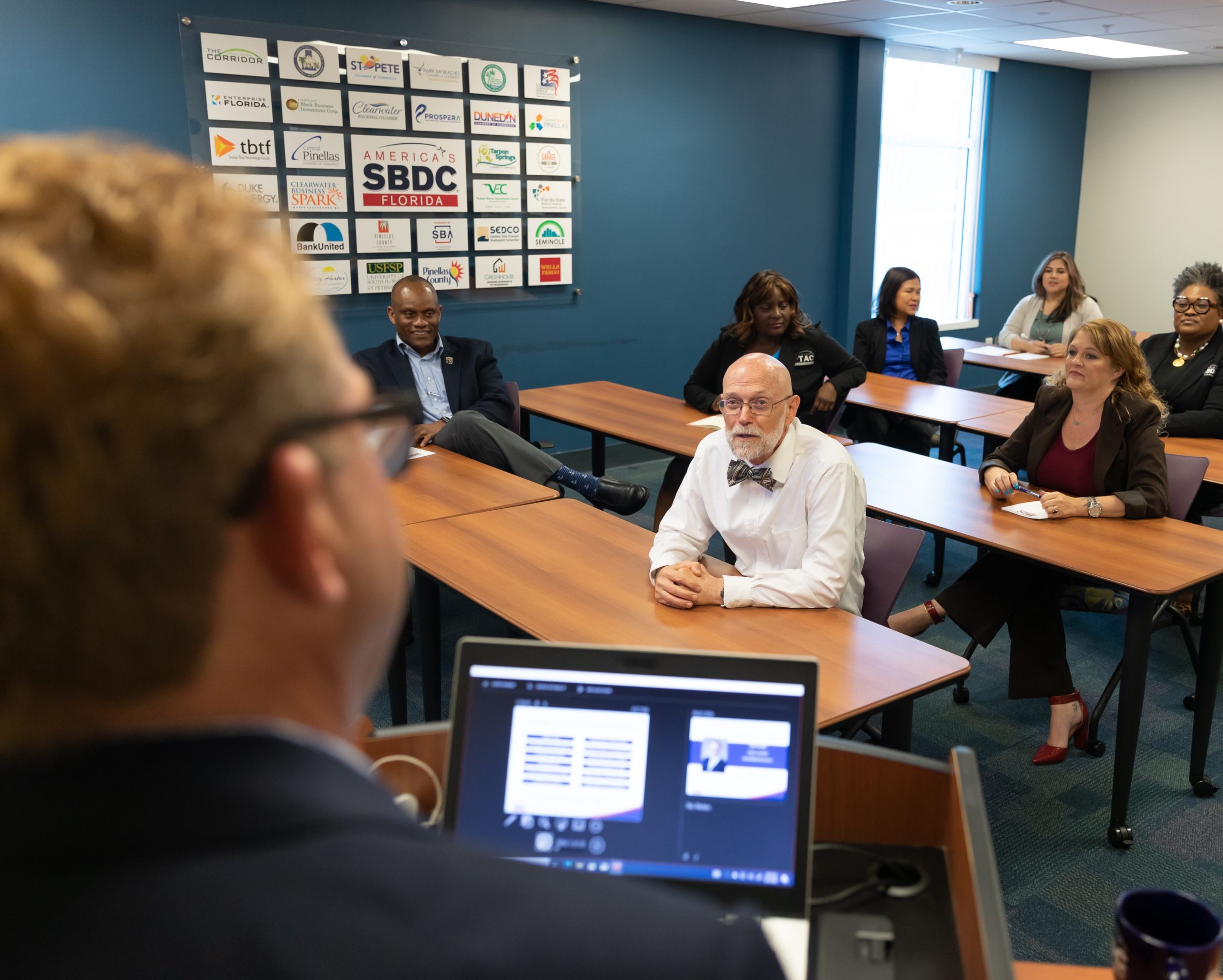 An image of a man leading a small business workshop in a meeting room with attendees at conference tables giving attention to the presenter.