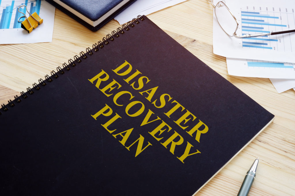 Image of a notebook with the title Disaster Recovery Plan on the front, located on a table with a pen and paper clip and eyeglasses.