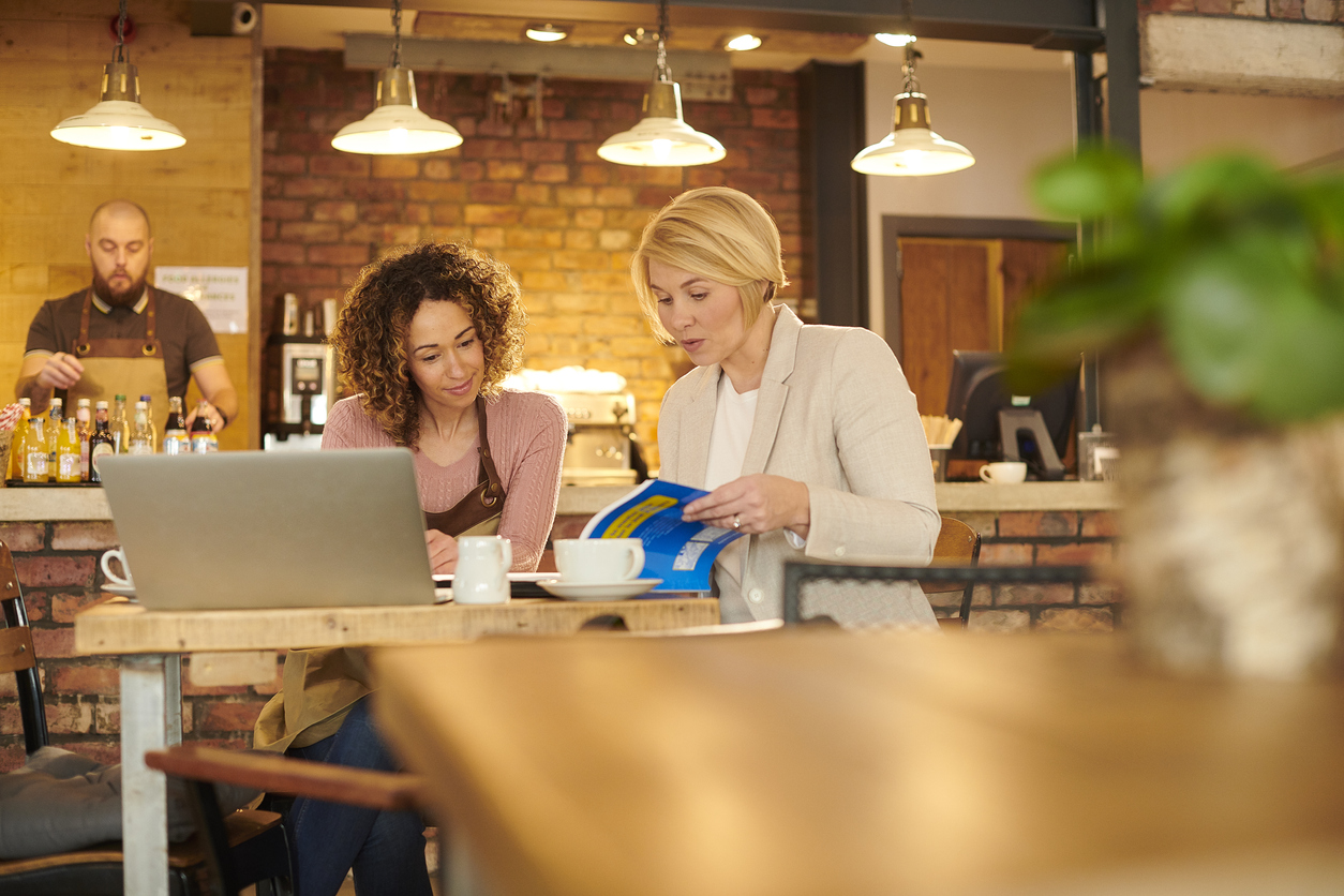 Image of two women meeting at a table in a coffee shop reviewing a book for small business advice, and an open laptop is on the table.