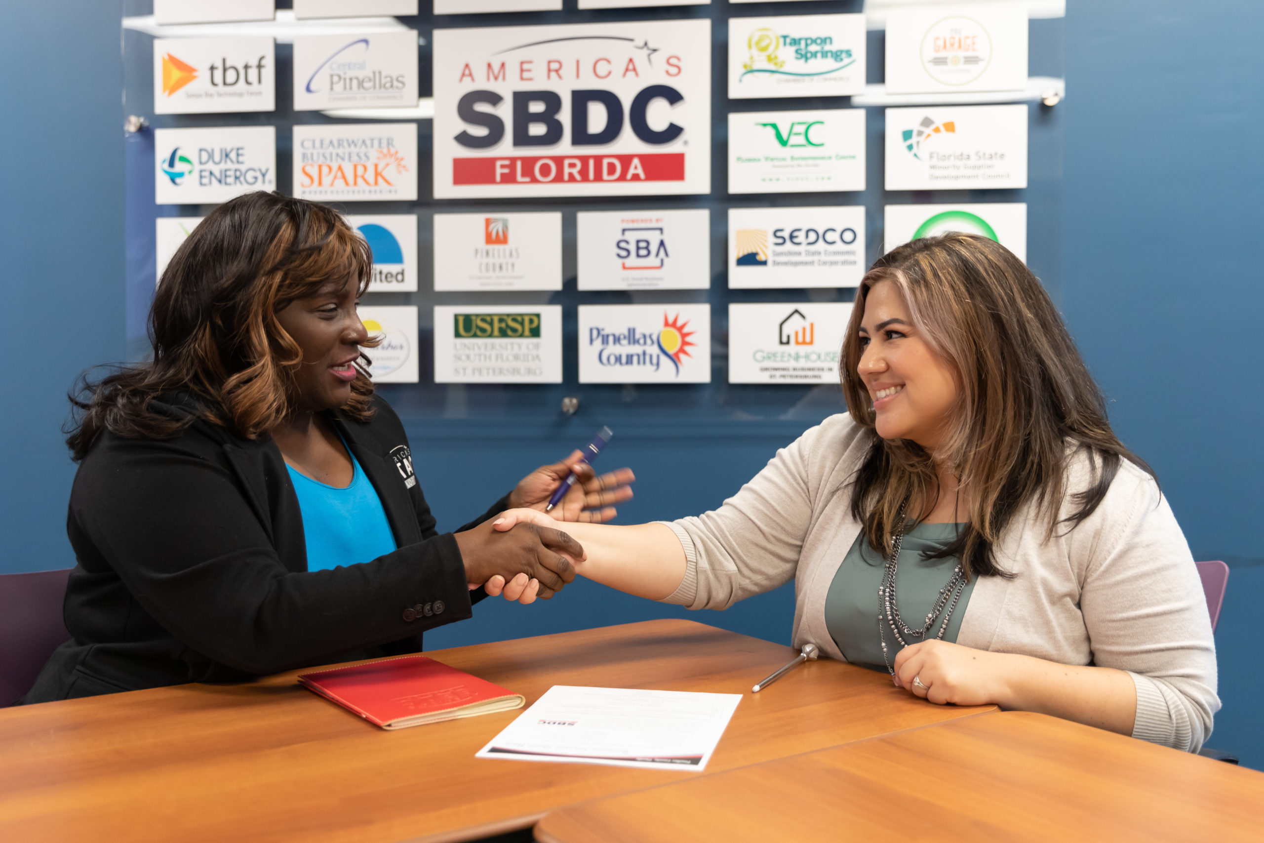 Two people shaking hands at a table with SBDC signage behind them.