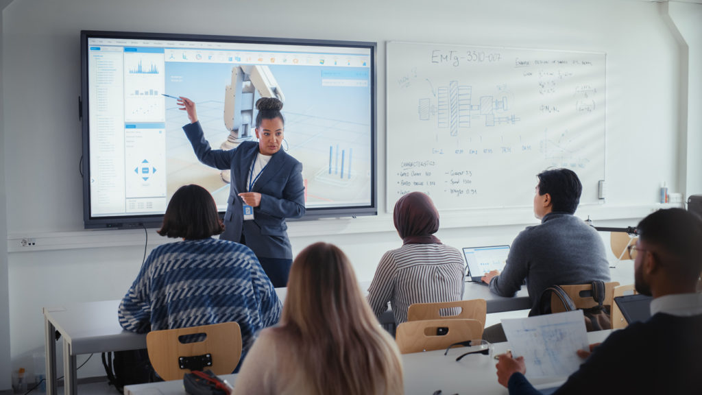 Image of University Robotics Lecture: Black Teacher Explain Engineering to Students. She Uses Interactive Whiteboard. Diverse Group of Young Engineers Listen in Class About Program Development for Robotic Arm.