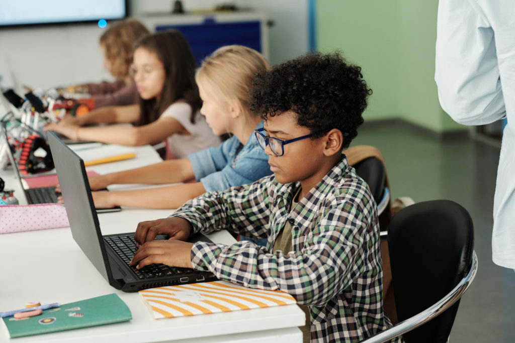 Image of side view of youthful African American schoolboy and his classmates working in front of laptops while sitting in row by desk.