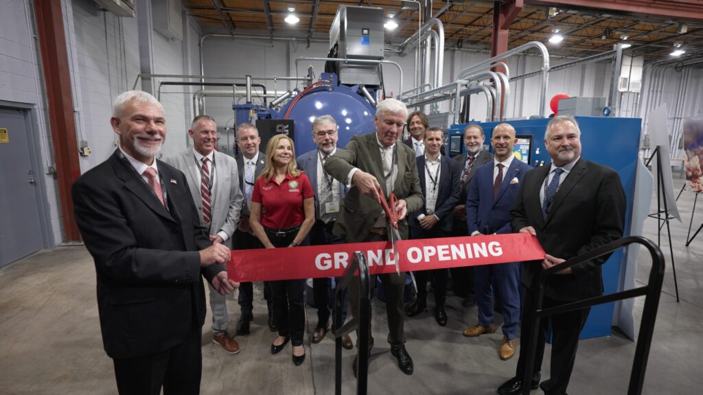 Image of group of people standing in front of machinery, one using scissors to cut a ribbon with "Grand Opening" printed on it